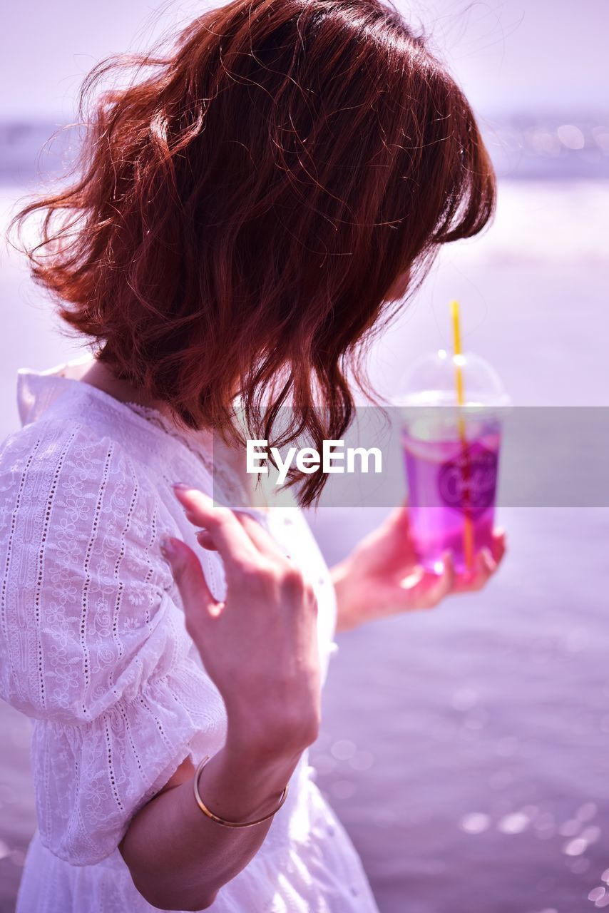 Side view of woman holding drink while standing against sea