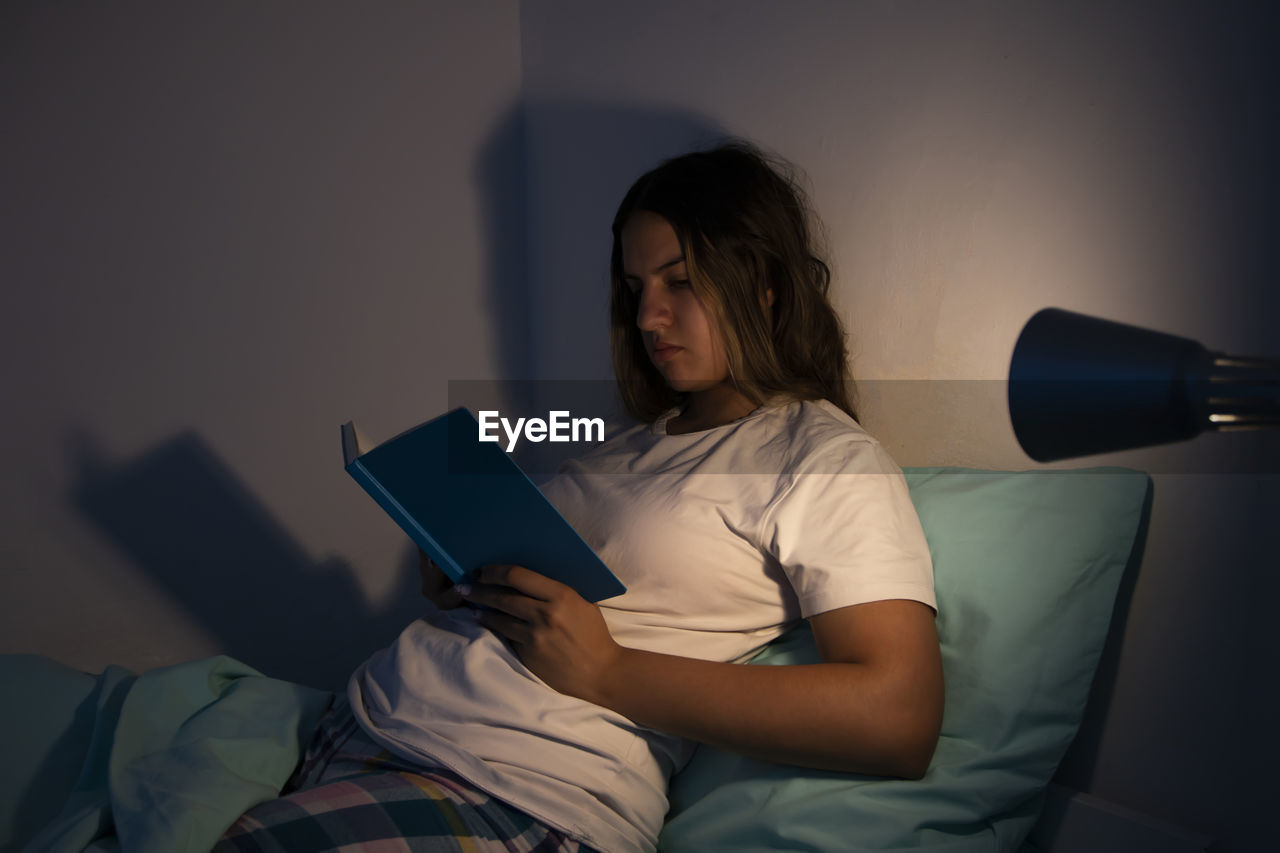 Teen girl lying on her bed, reading some notes at night with the illumination of her bedside lamp. 