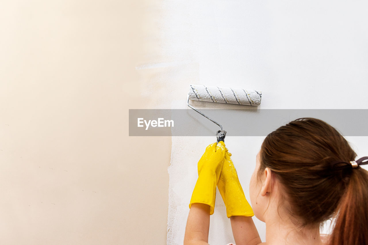 Teenage girl paints a wall with a roller of white paint, in yellow gloves