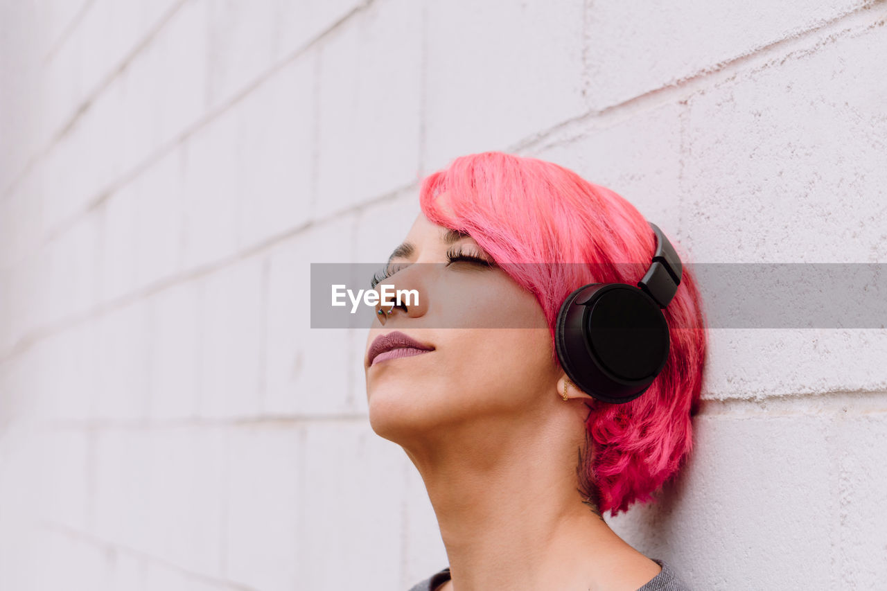Young female with bright pink hair listening to music with headphones while standing near white wall with eyes closed