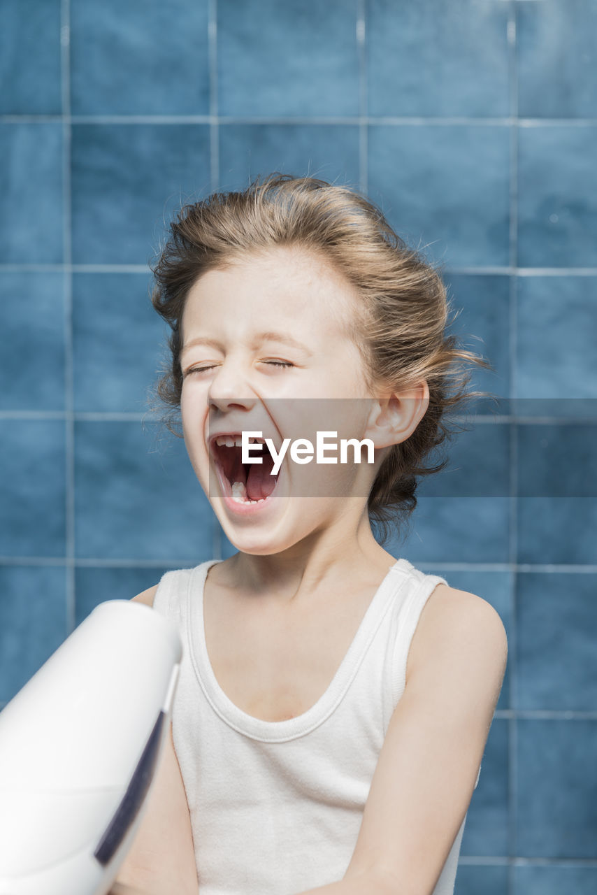 Cute boy with mouth open holding dryer against wall