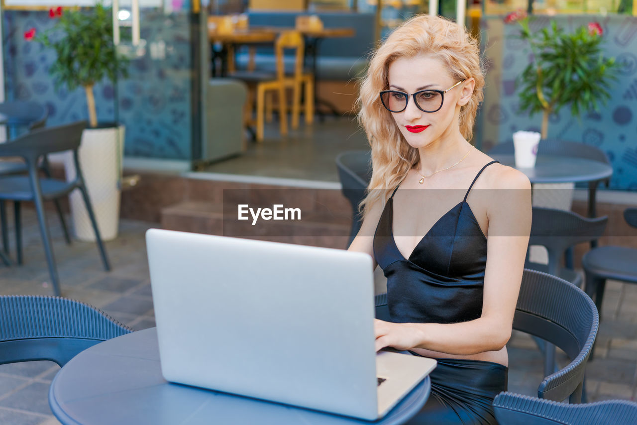 Beautiful woman in glasses sits at table in street cafe and works on a laptop.