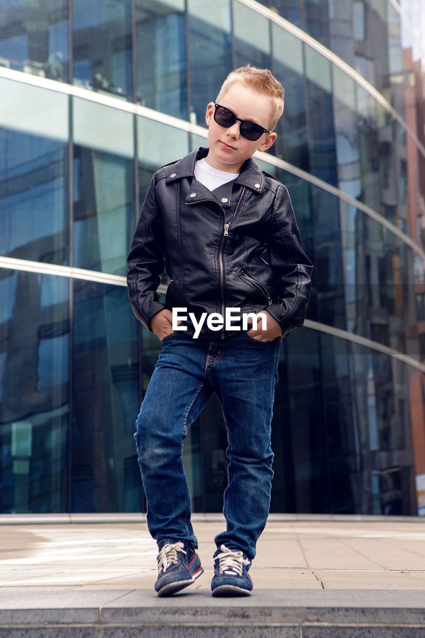 Baby boy in sunglasses and black leather jacket smiling on the background of the glass building