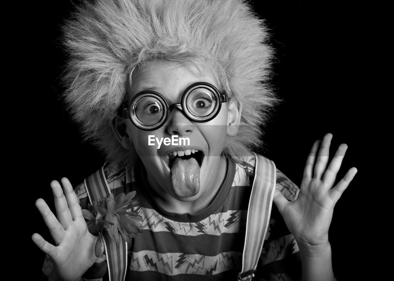 Portrait of funny boy sticking out tongue while wearing eyeglasses and wig against black background