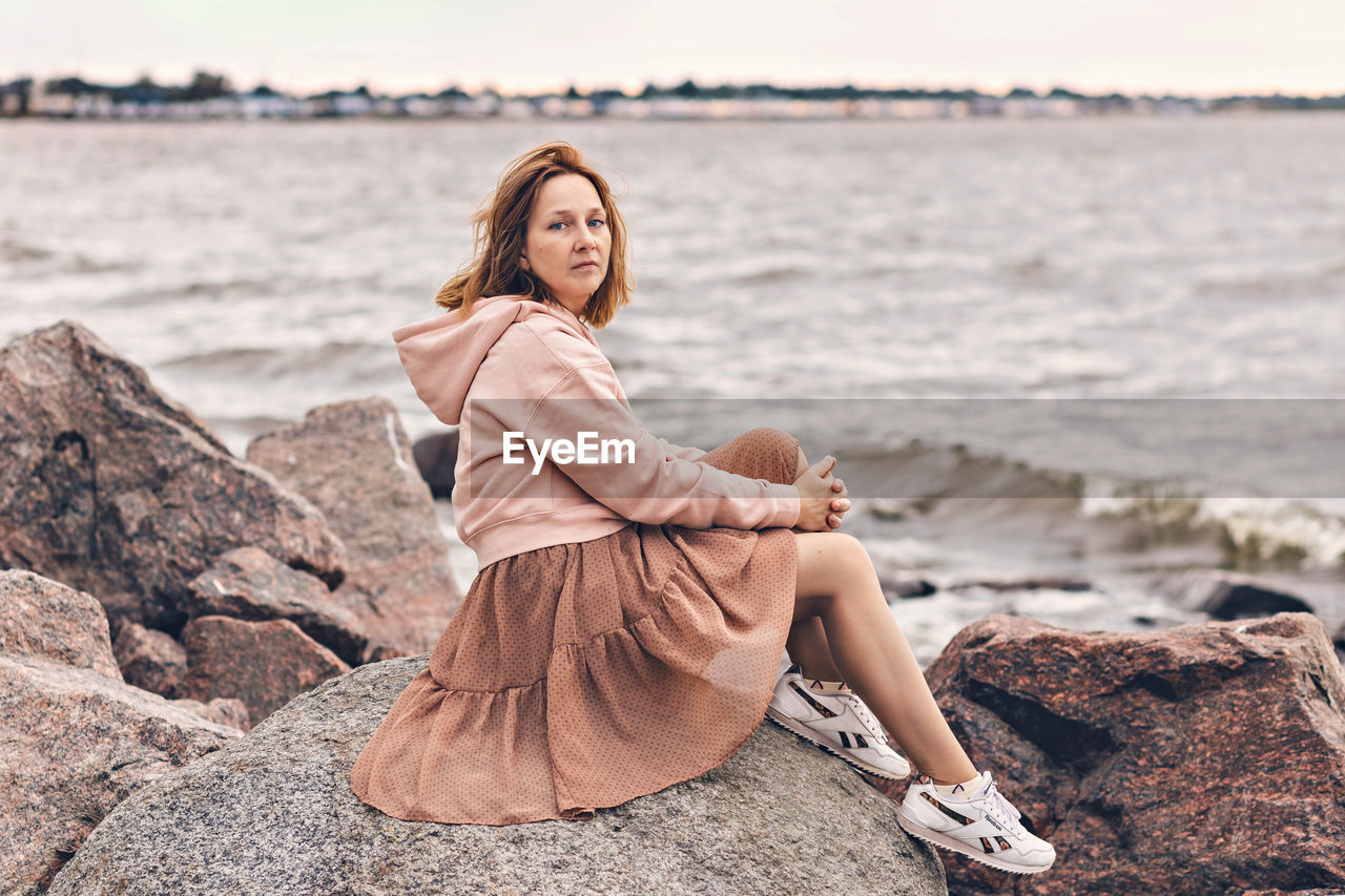 FULL LENGTH OF WOMAN SITTING ON ROCK AT BEACH