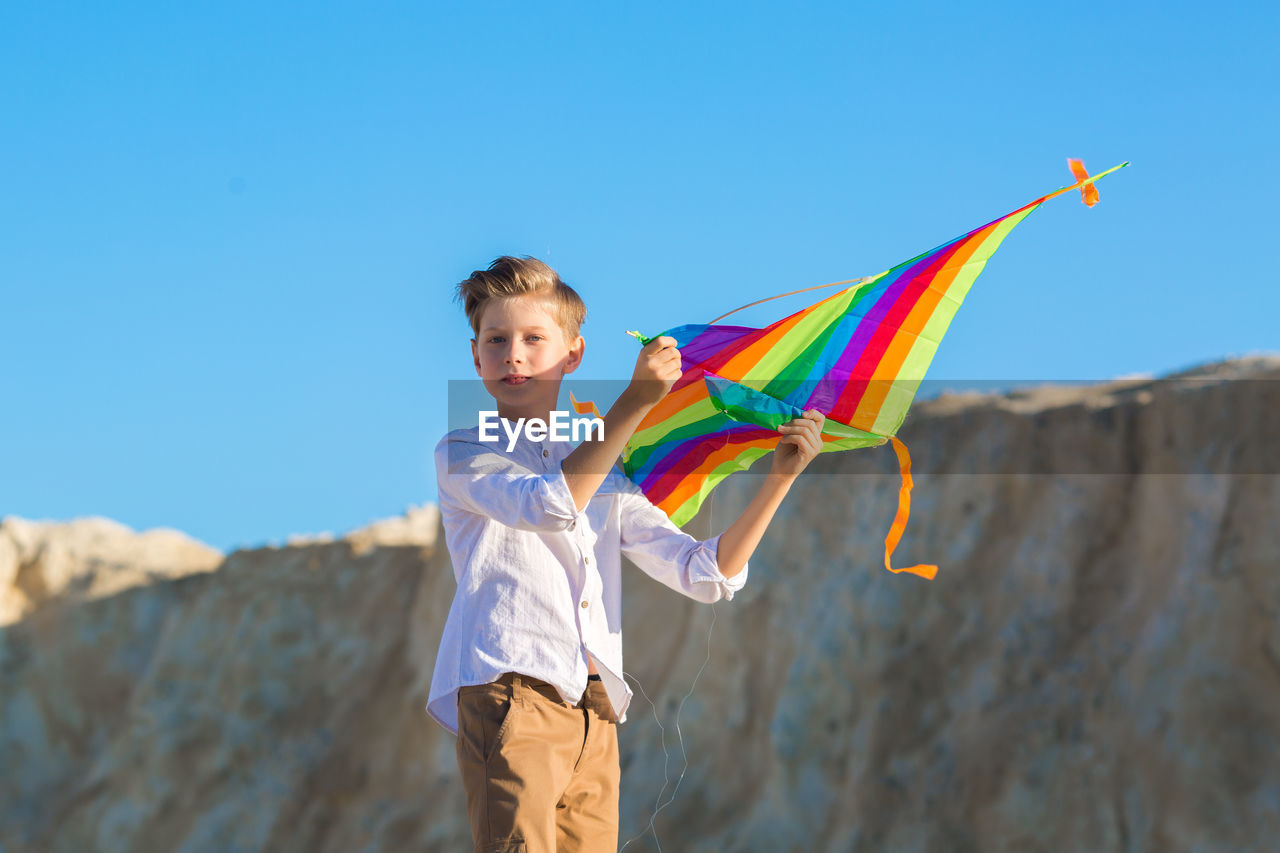 A handsome boy in a white shirt with a multicolored kite on blue sky background.