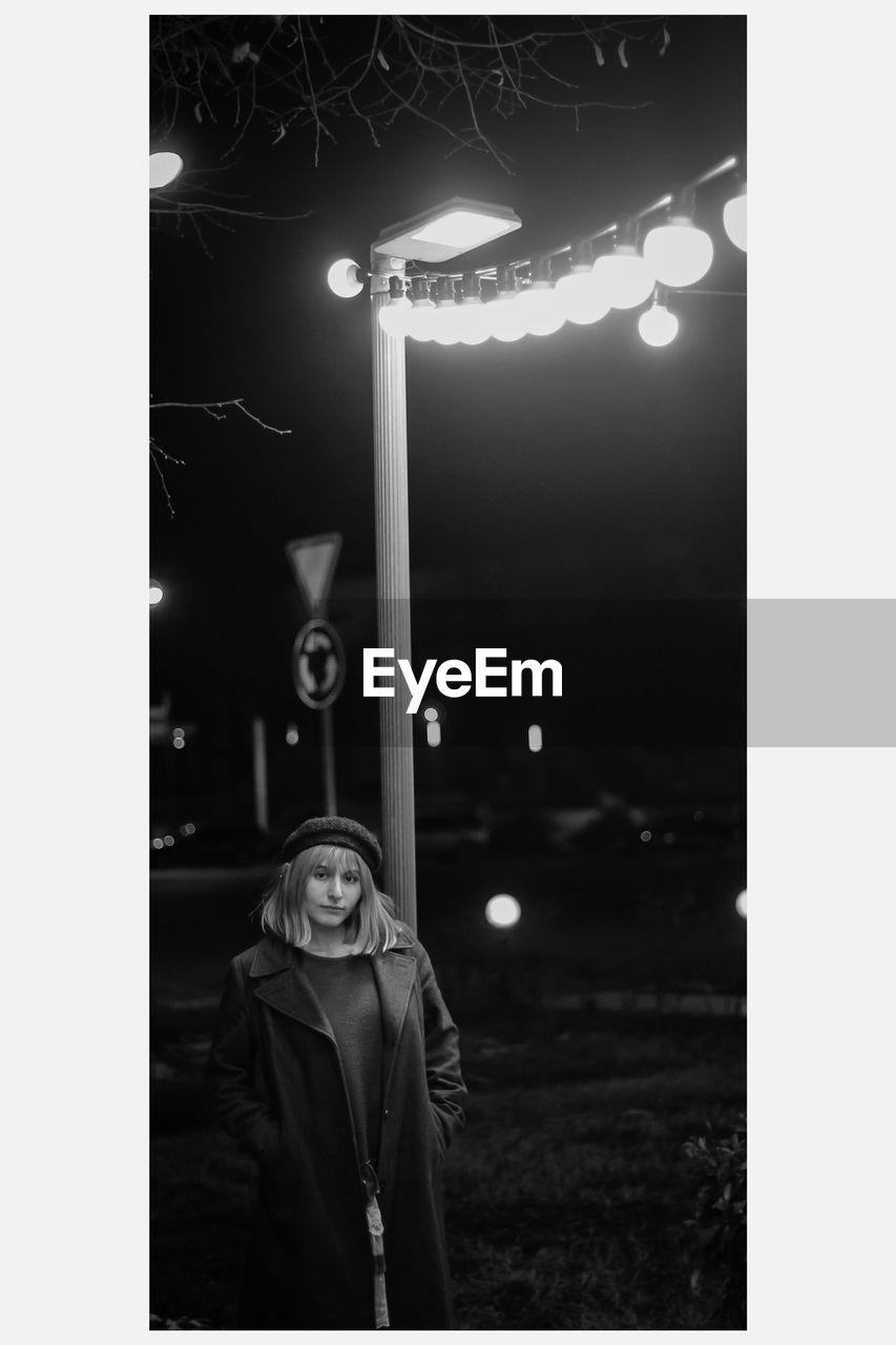 FULL LENGTH PORTRAIT OF WOMAN STANDING AGAINST ILLUMINATED LIGHTS