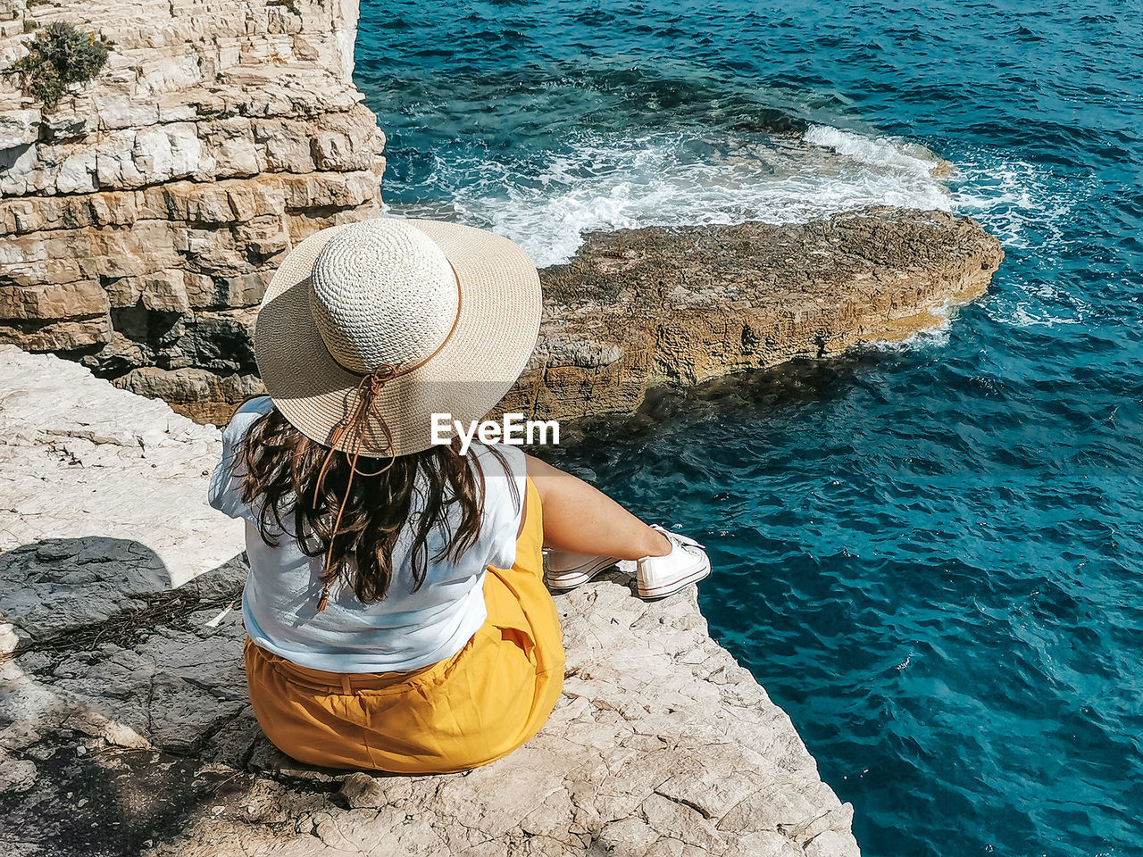 Young woman in yellow skirt sitting on edge of cliff above sea.