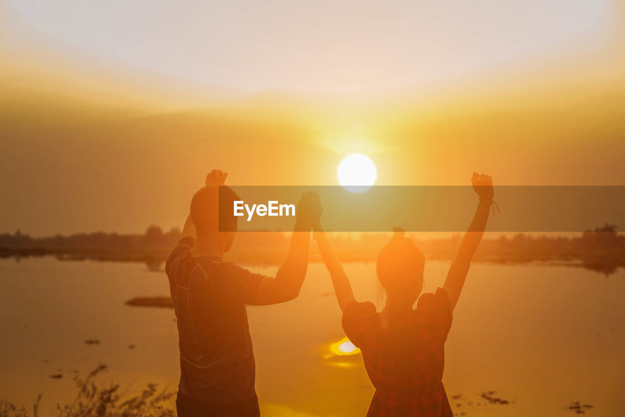 Rear view of couple holding hands while standing by lake against sky during sunset