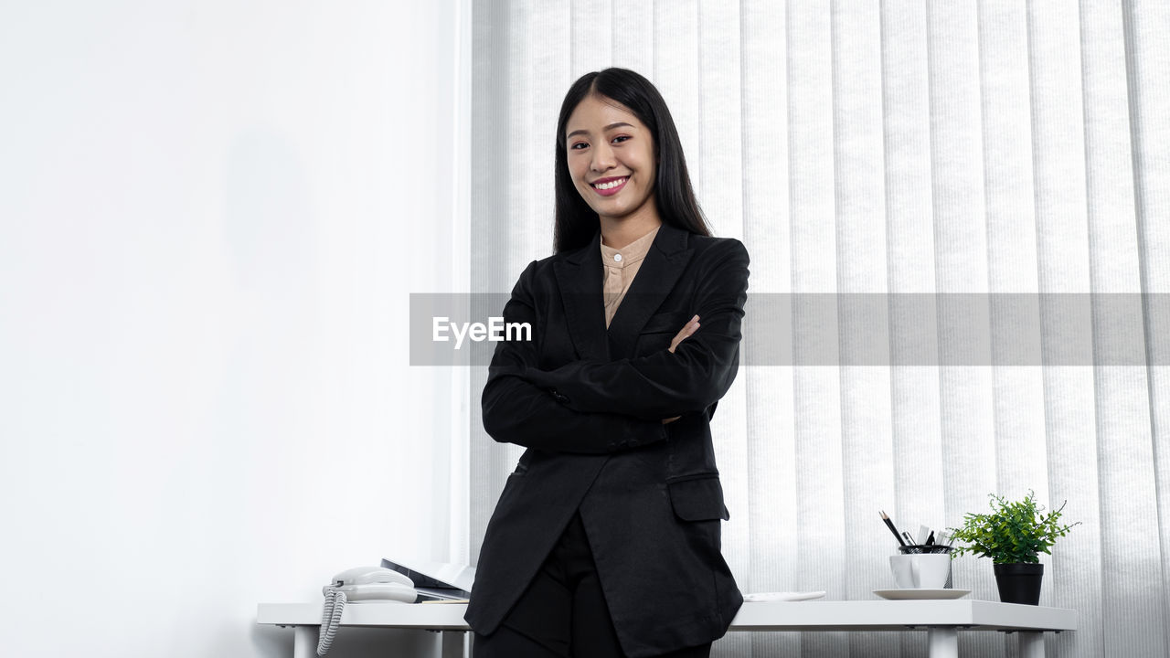 business, adult, one person, women, businesswoman, smiling, indoors, office, corporate business, happiness, business finance and industry, white-collar worker, sitting, formal wear, clothing, young adult, standing, emotion, table, female, businesswear, occupation, formal businesswear, portrait, three quarter length, looking at camera, furniture, working, success, cheerful, outerwear, person, businessman, professional occupation, arms crossed, black hair, front view, copy space, waist up, hairstyle, houseplant, black, long hair, communication, conversation