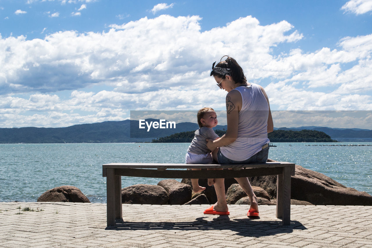Full length of mother sitting with son on bench at promenade by sea against sky