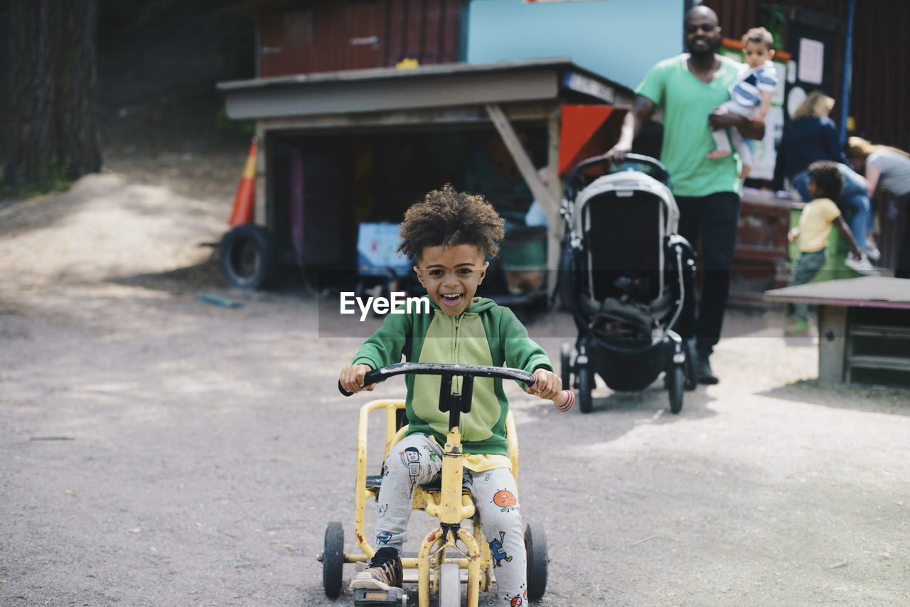 Happy boy riding tricycle while father and brother walking in background