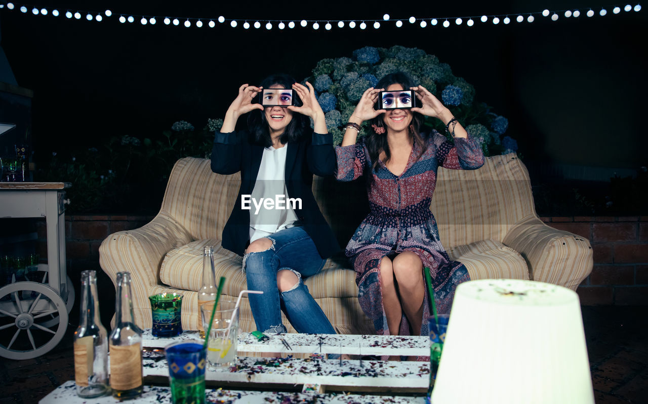 Portrait of young friends holding smart phones on eyes while on sofa during party