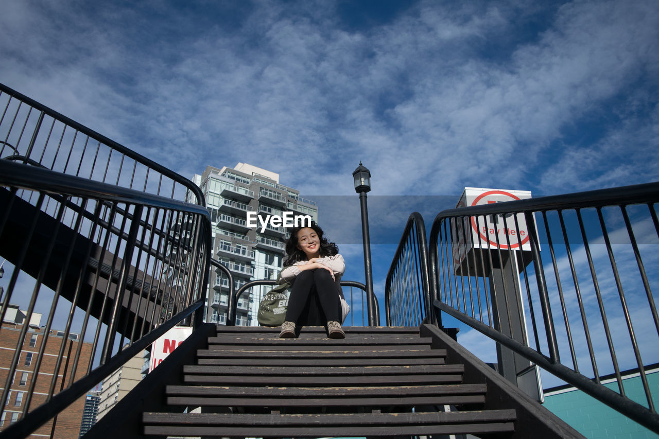 Low angle portrait of smiling young woman sitting on steps against sky