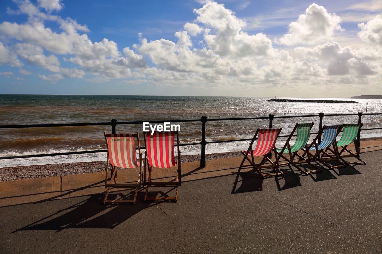 Empty lounge chairs on promenade by sea against cloudy sky