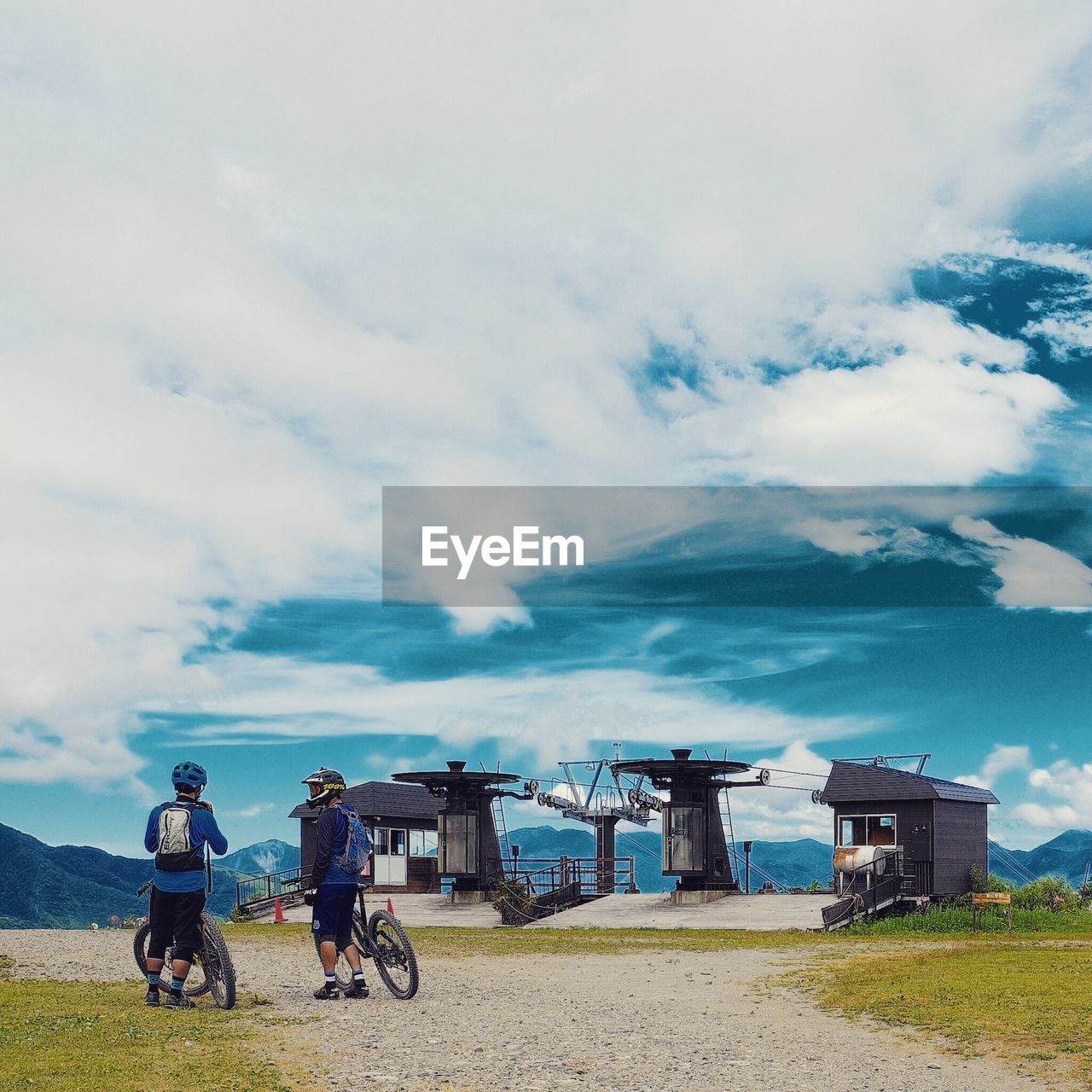 Rear view of bikers with bicycles on dirt road against cloudy sky