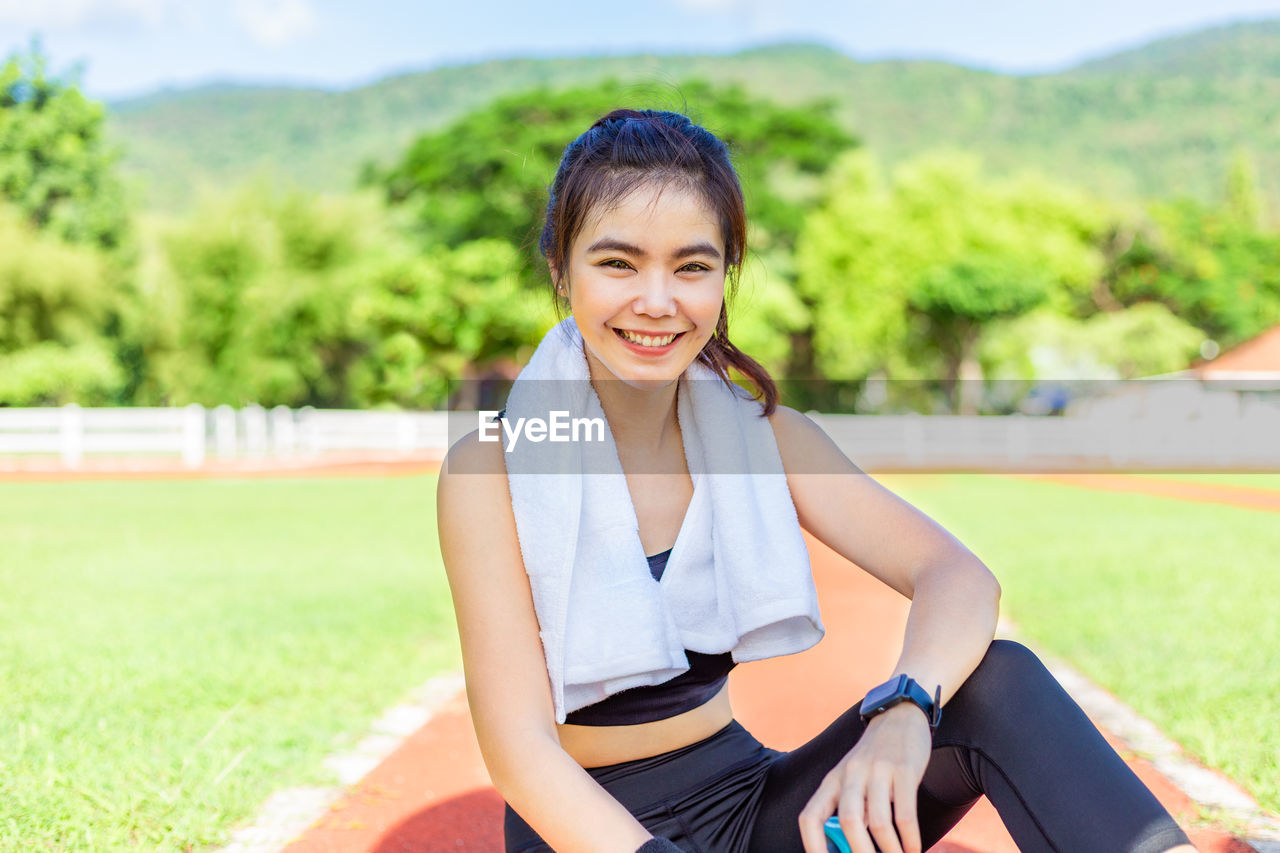 Beautiful young happy asian female athlete sitting on running track smiling at camera on sunny day
