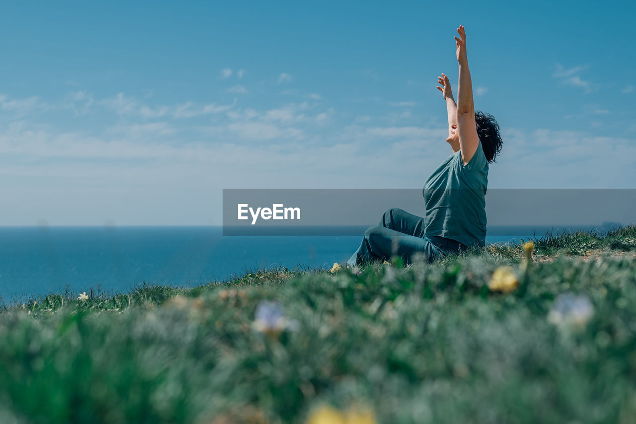 An adult woman sits on a mountain barefoot in a lotus position, raising her hands up to the sky and