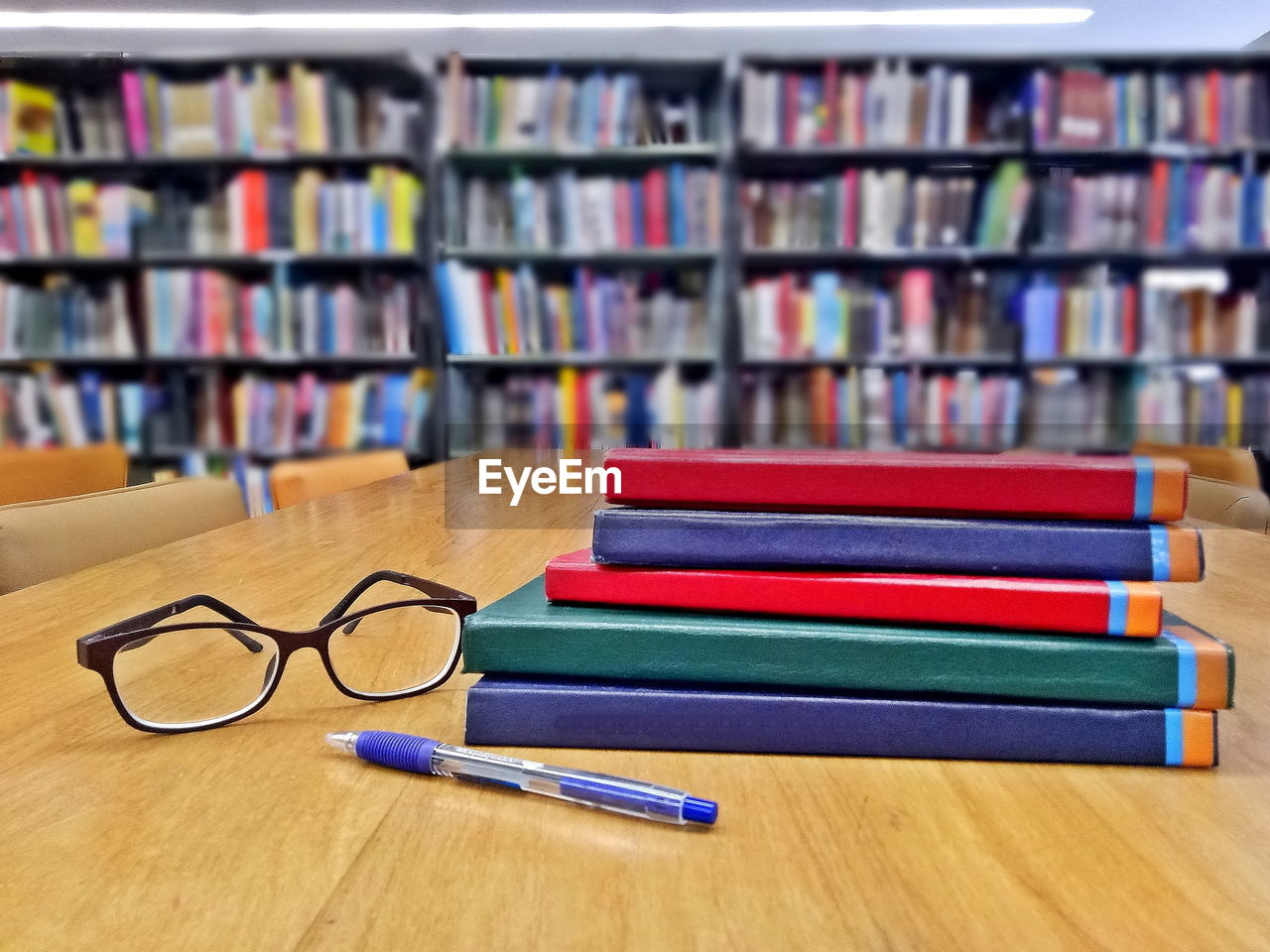Glasses, pen and books on wooden table in library with bookshelves background.