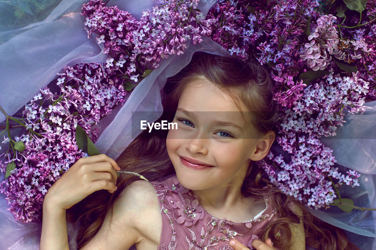 Child girl in a purple floral dress lies on the ground among lilacs on a veil in spring