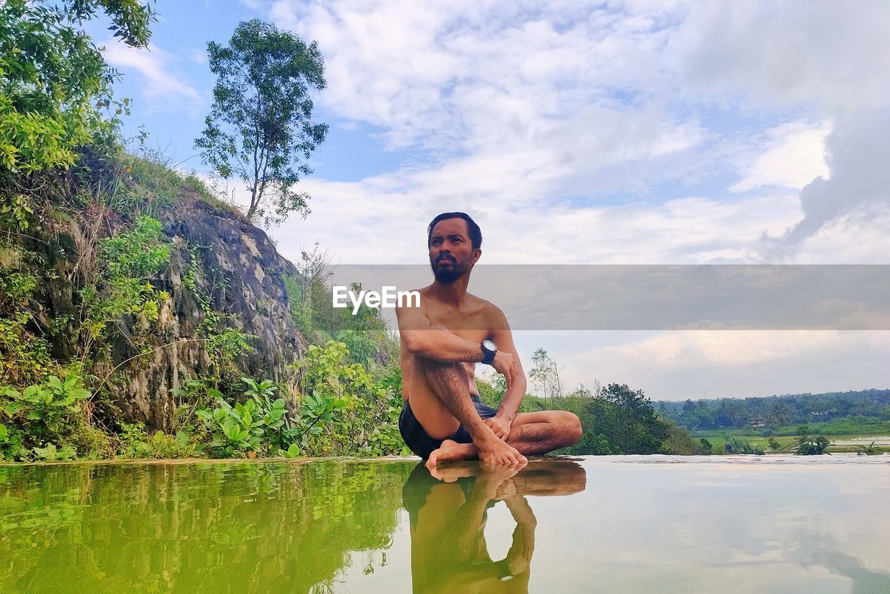 Full length of shirtless man standing by lake against sky