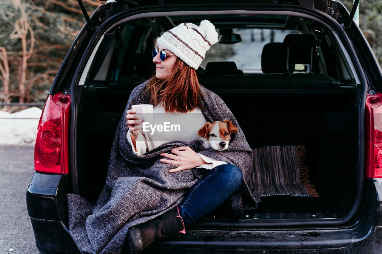 Woman holding mug sitting with dog in car trunk