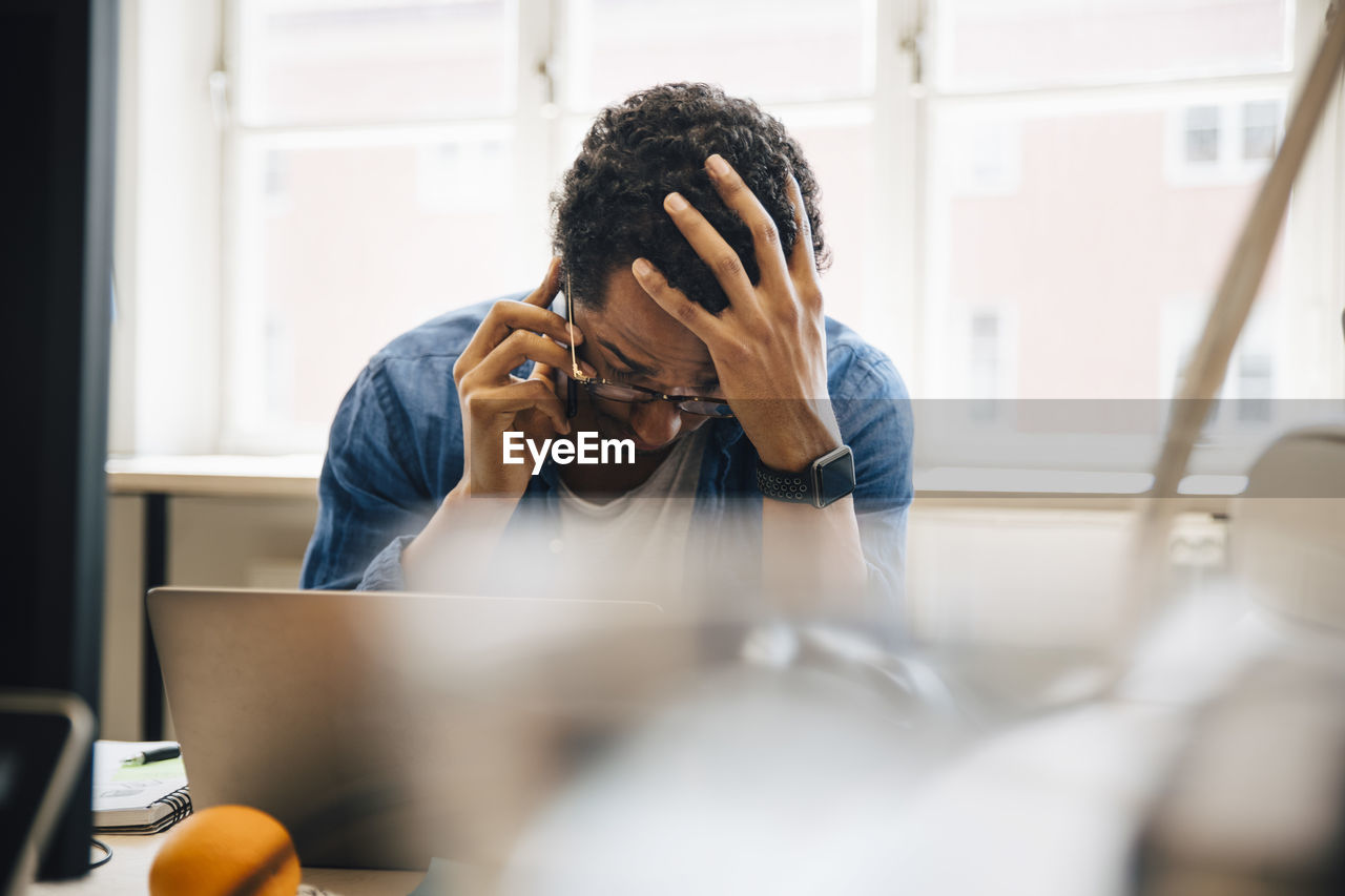 Frustrated male computer hacker with hand in hair talking on smart phone while using laptop in creative office