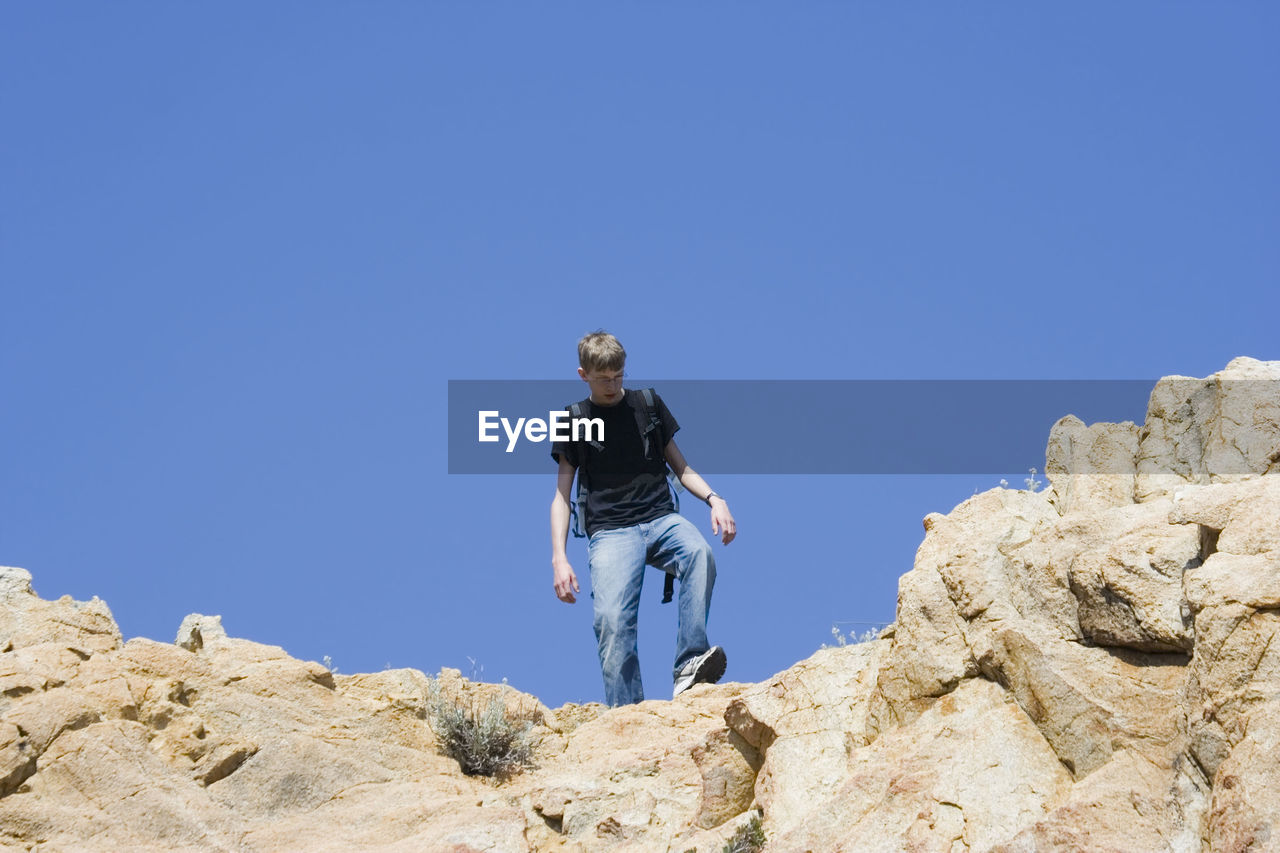 Low angle view of male hiker moving down on rocks against clear blue sky