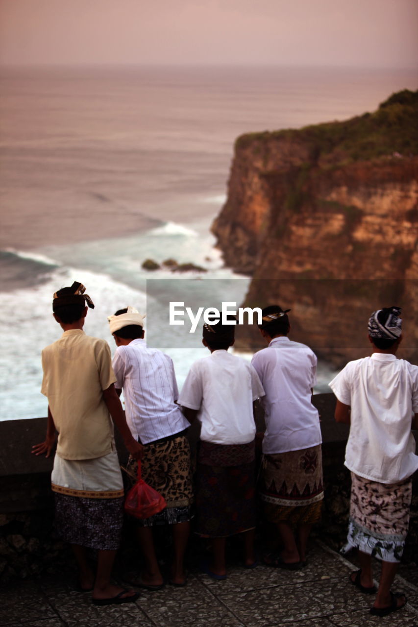 Rear view of boys wearing headdresses standing by retaining wall against sea during sunset