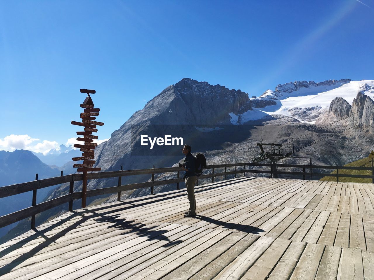 Side view of man reading sign board at observation point by rocky mountain against blue sky