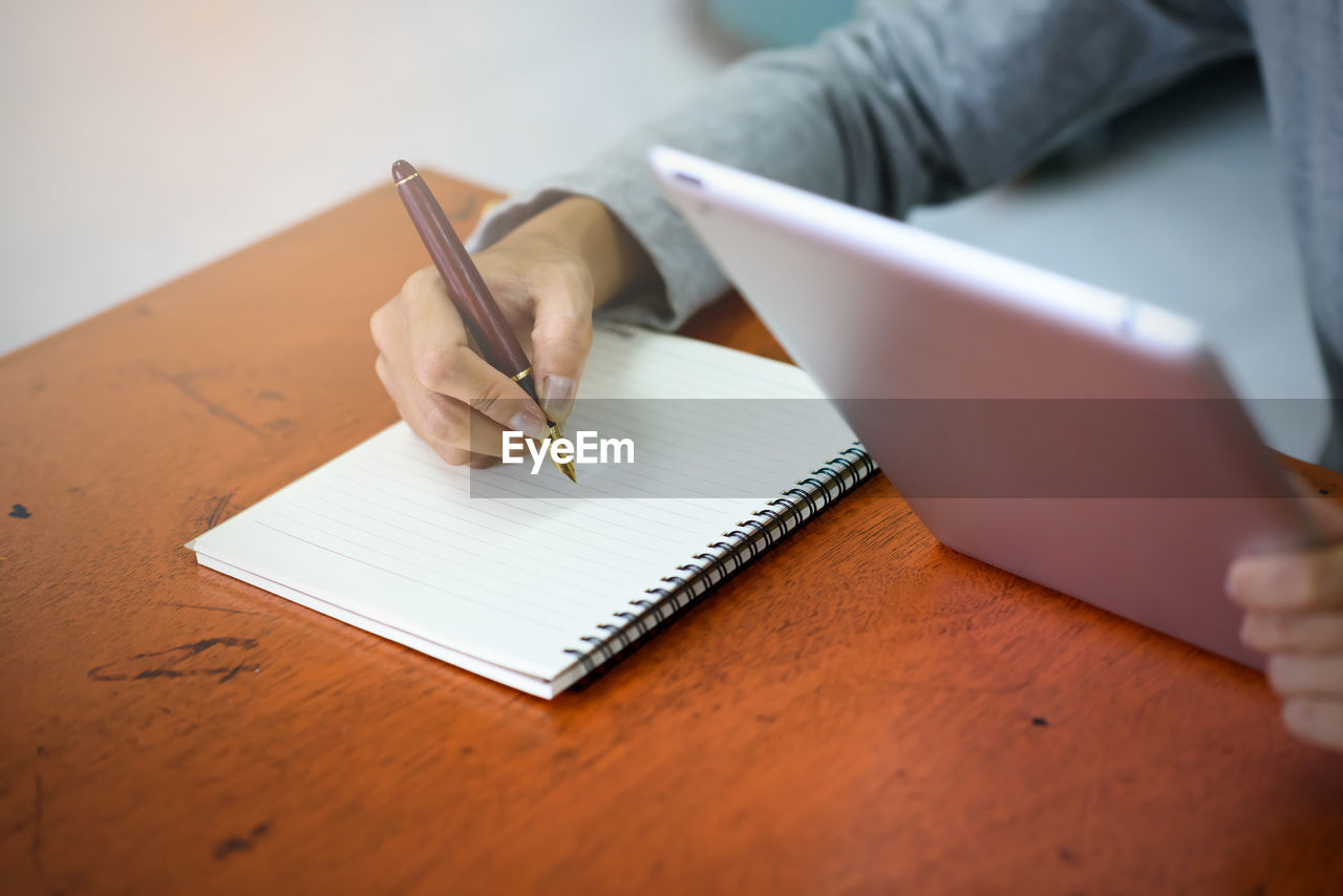 Close-up of businesswoman holding digital tablet while writing on book in office