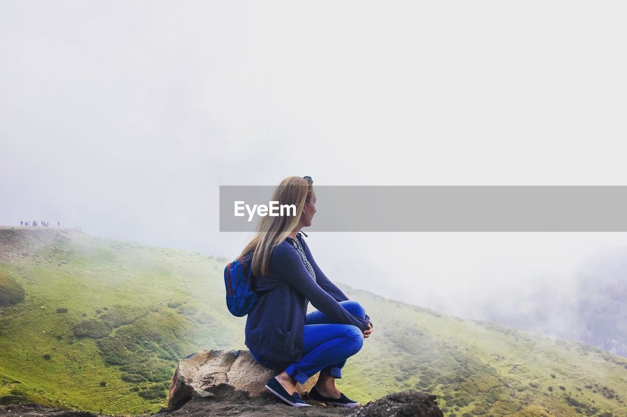 Side view of young woman sitting on rock and looking at view during foggy weather