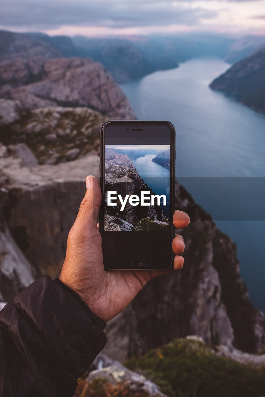 Hand holding smartphone with image of scene in background on it at norwegian fjords.