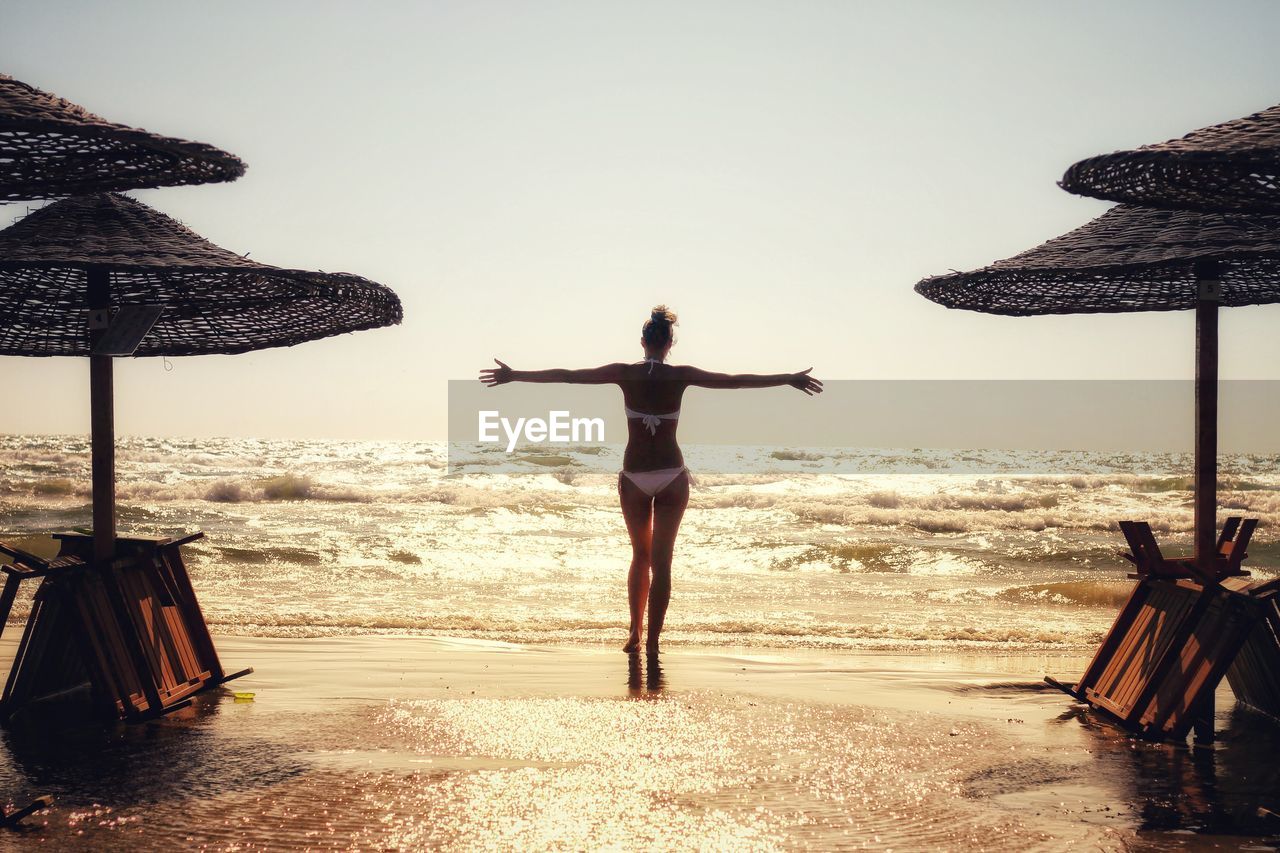 Rear view of beautiful woman standing on beach against clear sky at sunset