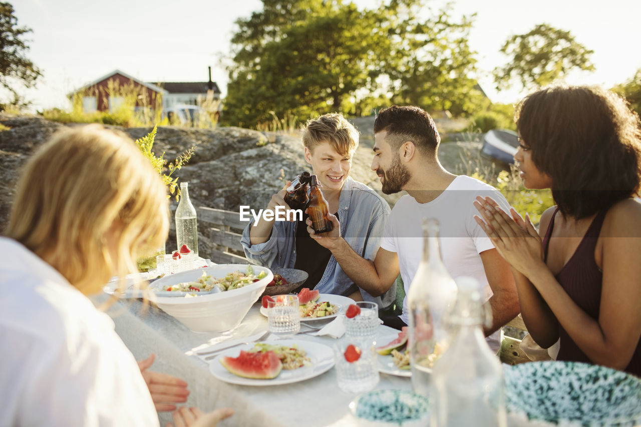 Men toasting beer bottles while sitting with friends at picnic table