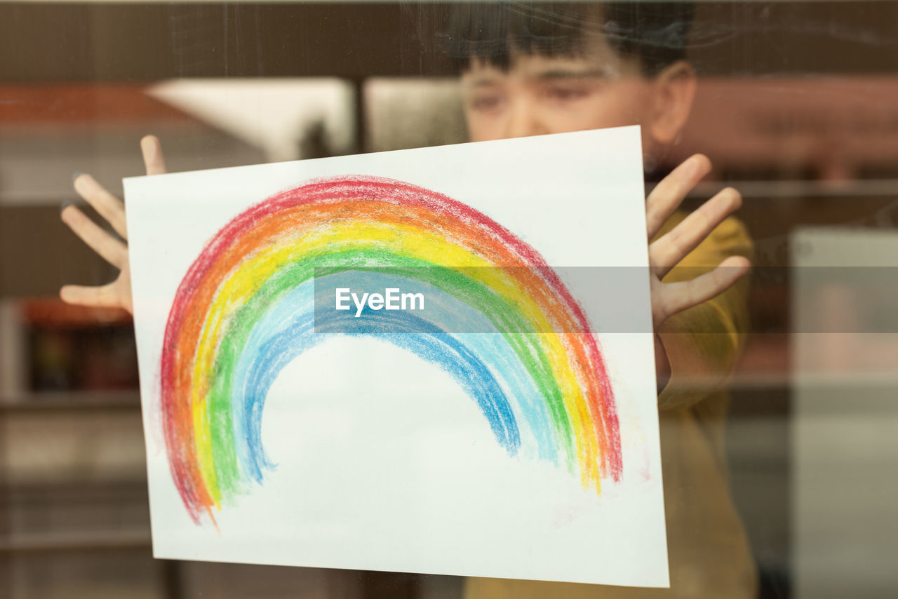 Boy holding multi colored rainbow painting seen through window