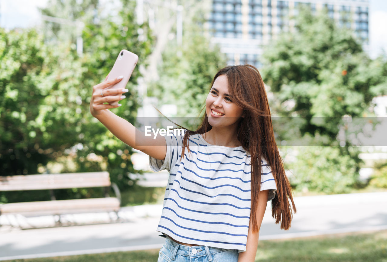 Portrait of young woman student with long hair taking selfie with mobile phone in city park