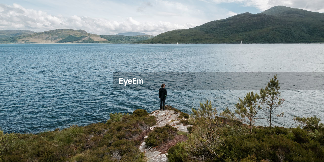 REAR VIEW OF MAN STANDING BY SEA AGAINST SKY