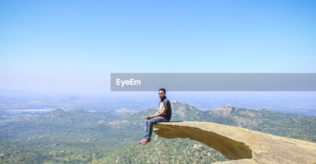 Man sitting at the edge of cliff against clear blue sky