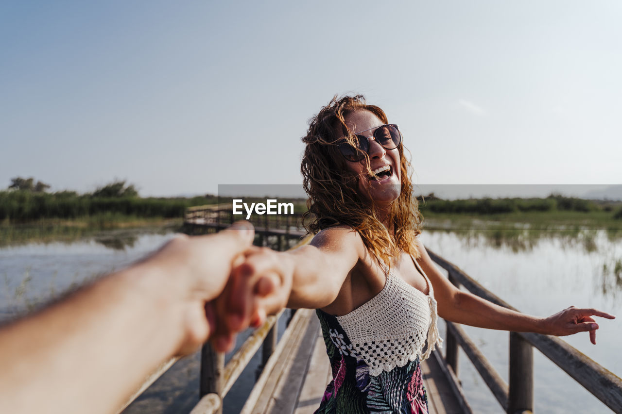 Woman holding hand of boyfriend while standing on footbridge against sky