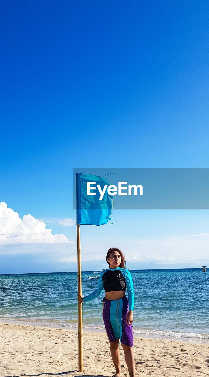 Woman standing by flag at beach against blue sky