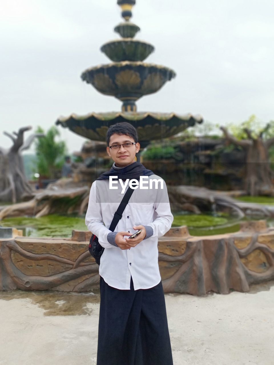 Portrait of young man standing against fountain and sky