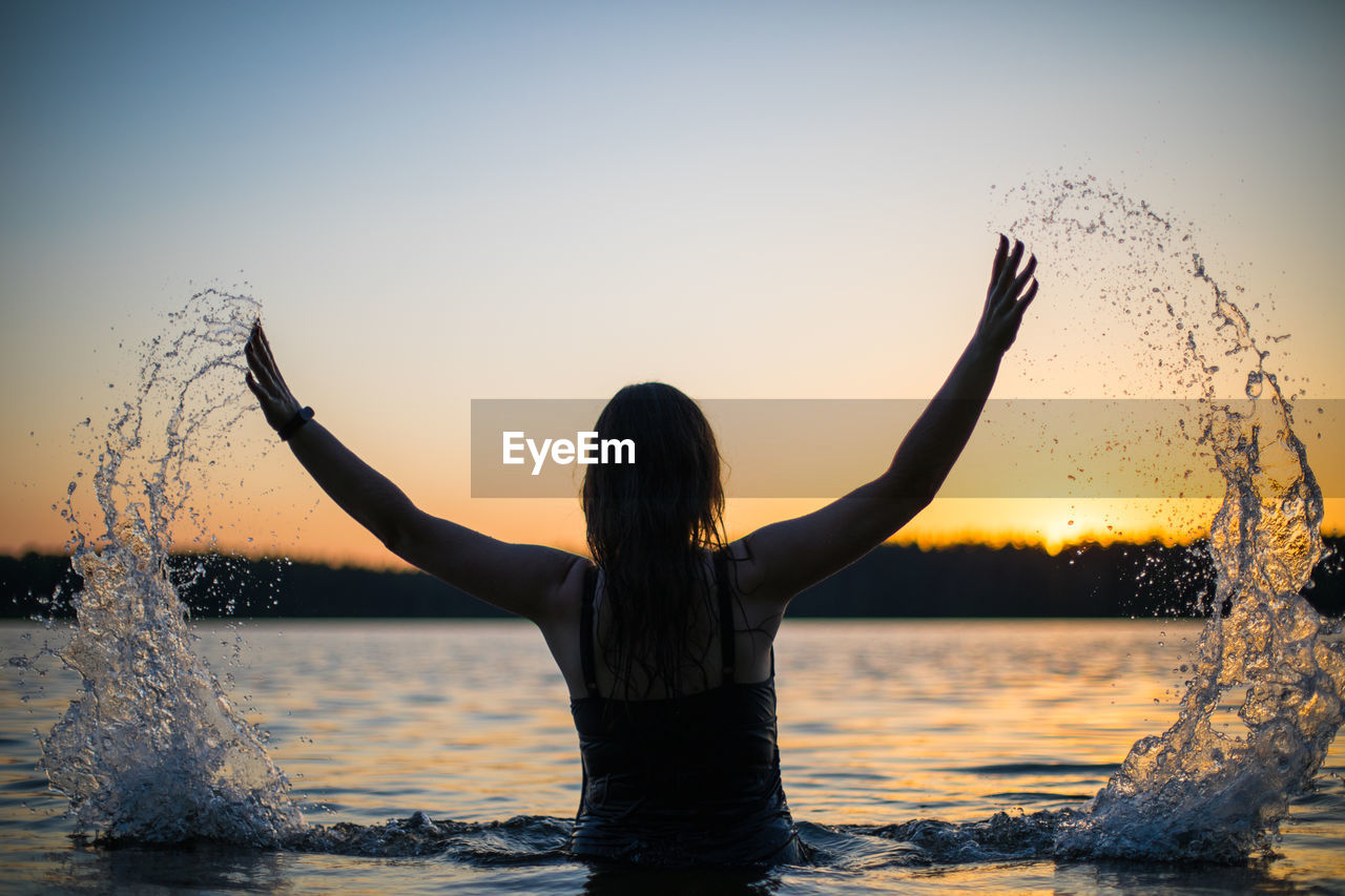 Girl in a long black swimsuit swims outside the city on the lake in the rays of sunset or dawn.