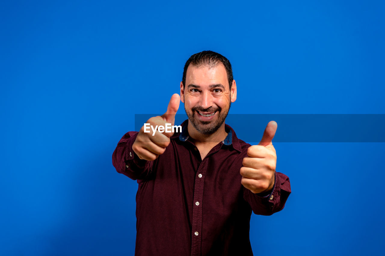 blue, sign language, one person, gesturing, men, studio shot, adult, thumbs up, emotion, portrait, smiling, happiness, colored background, looking at camera, blue background, hand sign, positive emotion, cheerful, hand, finger, front view, waist up, person, copy space, communication, indoors, pointing, fun, casual clothing, standing, showing, facial expression, young adult, excitement, success, cut out, facial hair