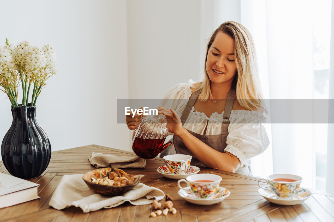 Blond woman pouring hibiscus tea in cups while sitting at table