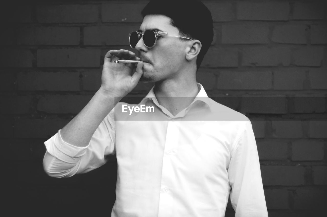 Young man smoking while standing against brick wall