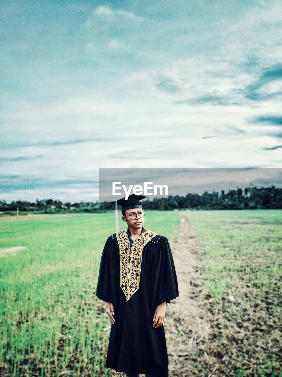 Young man wearing graduation gown while standing on grassy field against sky