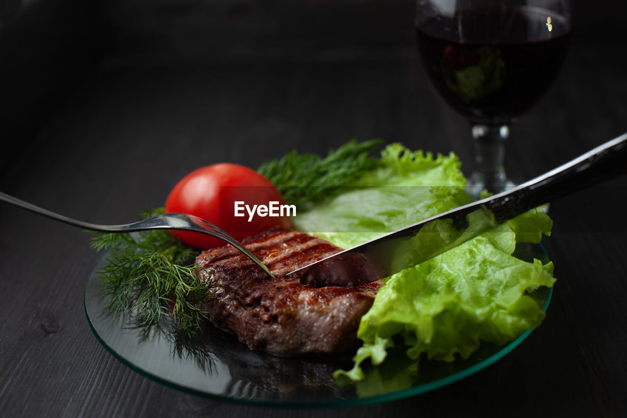 A delicious slice of steak on a plate with lettuce and tomato. knife and fork are stuck in the meat.