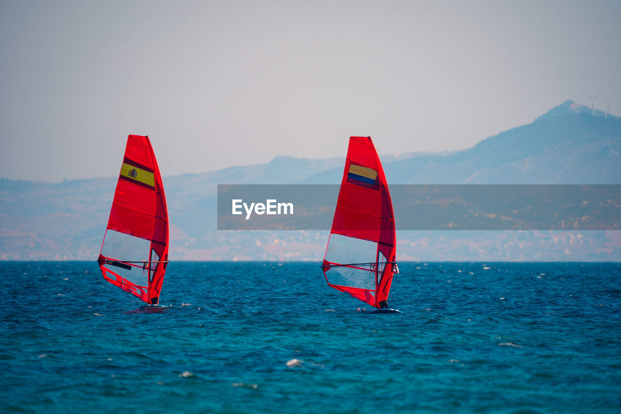 Red sails of windsurfers on rippling blue water of sea bay with blurred mountains on background in sunlight