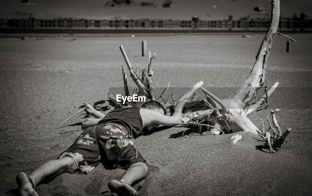 Full length of tired man sleeping by driftwood on sand